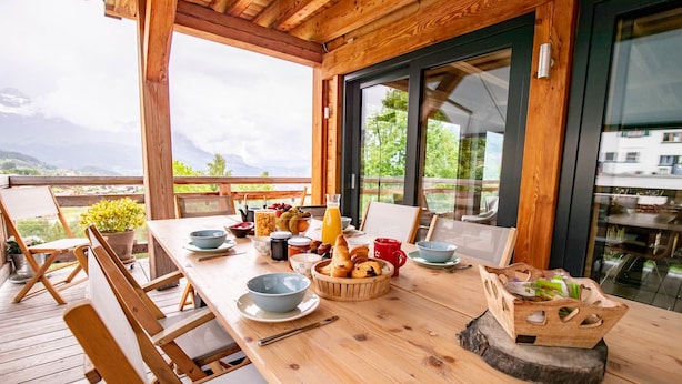 A wooden deck with a table set for breakfast, including bread, fruits, and beverages. - Chalet Caprice, 8 people (COMBLOUX)