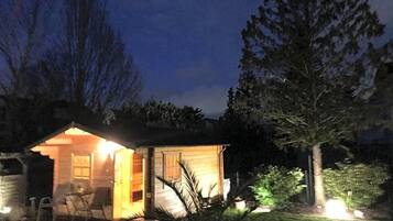 Eine kleine Holzhütte mit offener Tür, einer Terrasse mit Sitzgelegenheiten und einem gut beleuchteten Innenraum.