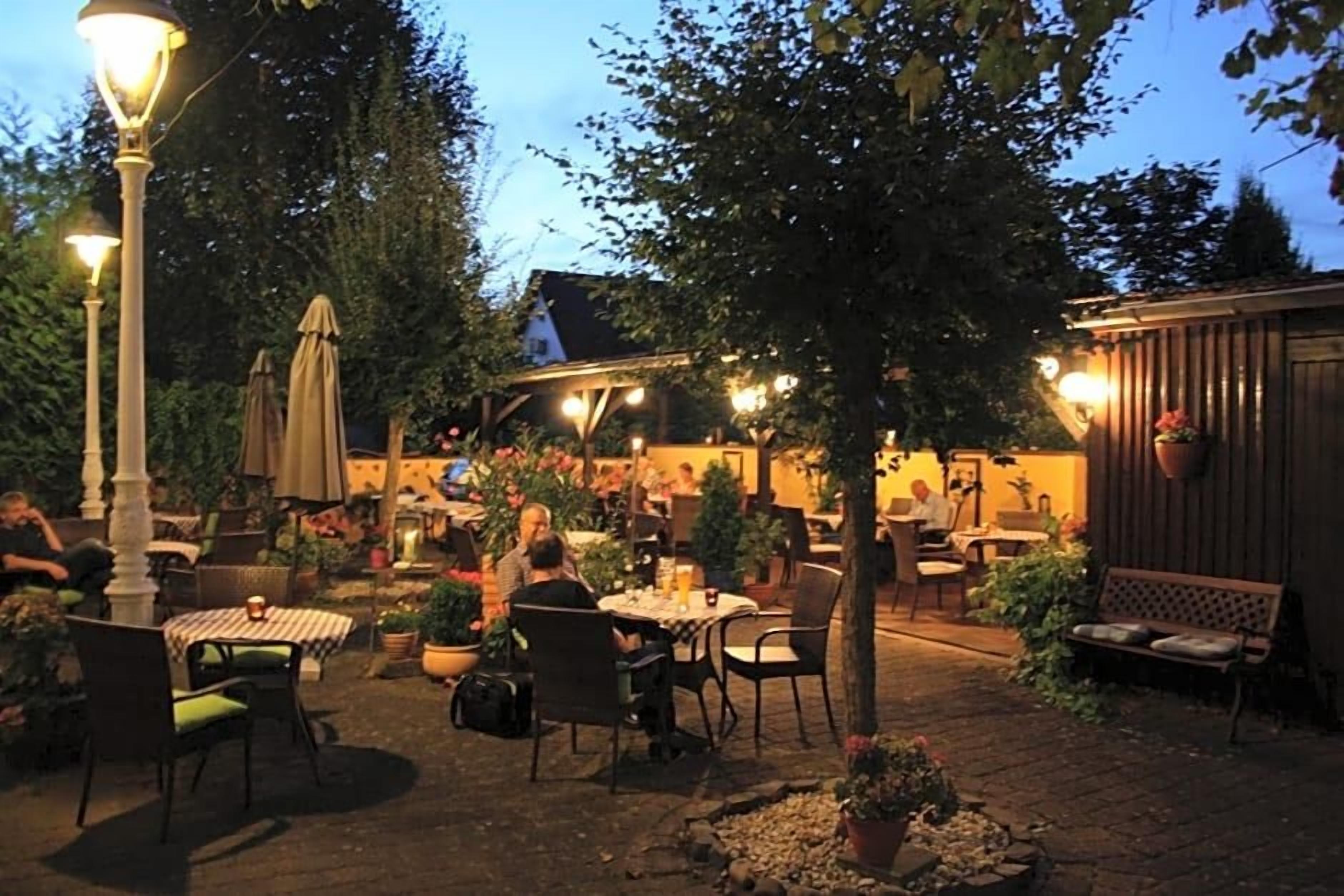 Area patio esterna con tavoli e sedie, circondata da piante in vaso e alberi.
