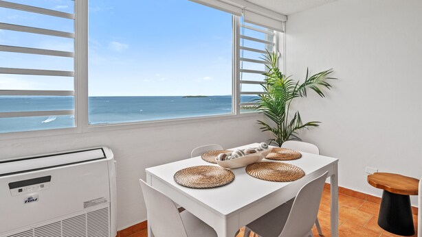 Una habitación con una mesa blanca, un ventilador de techo y vista al océano a través de amplias ventanas.
