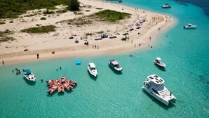 Vista aérea de una playa con aguas turquesas cristalinas, varias embarcaciones ancladas cerca de la orilla y una playa de arena con gente y sombrillas.