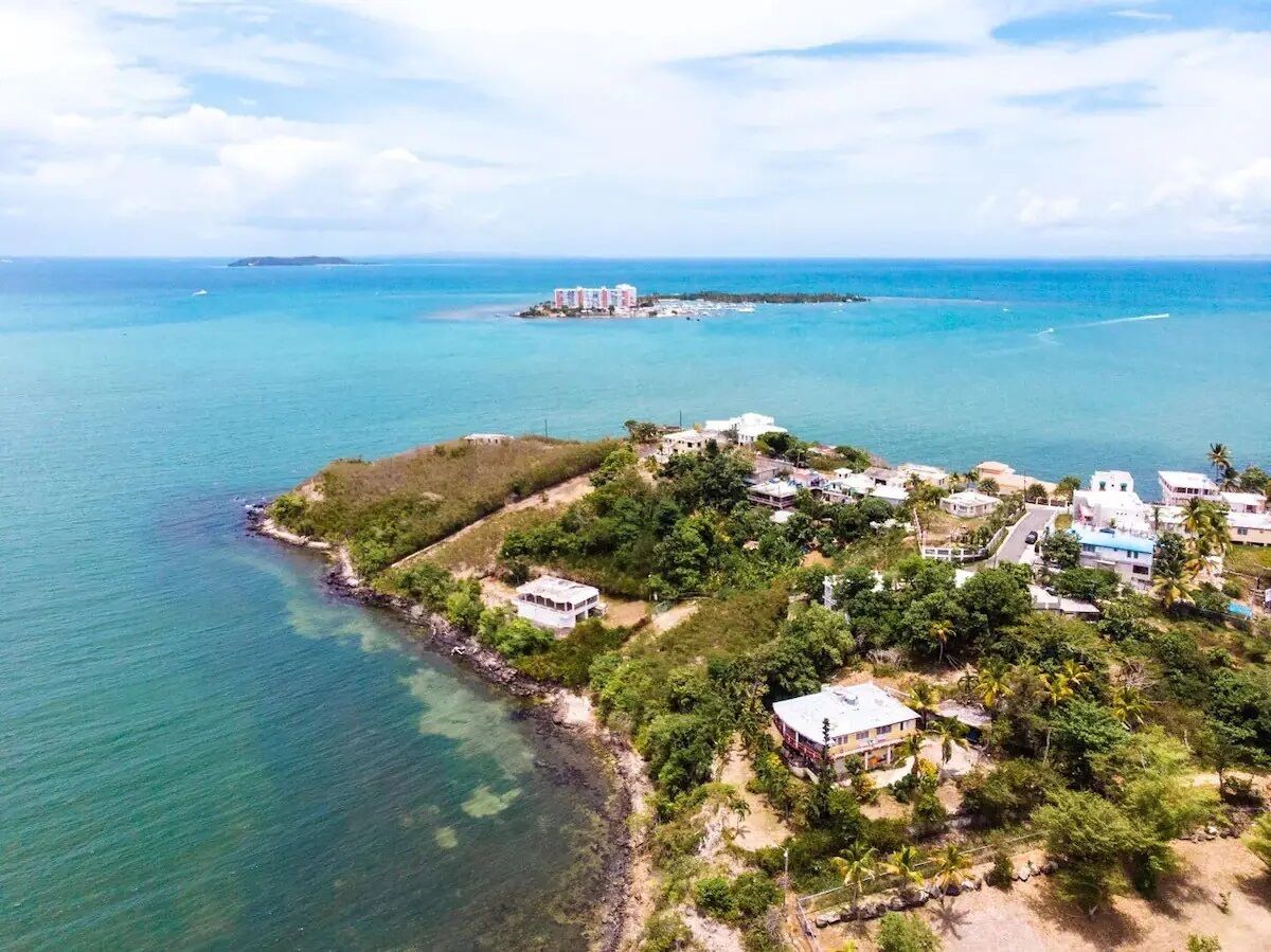 Une vue aérienne d’une zone côtière avec des bâtiments, des eaux turquoise et limpides, et une île au loin.