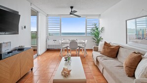 Una sala de estar moderna con un televisor de pantalla plana, un comedor con mesa y sillas, y un ventanal con vistas al mar.