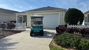 Une voiturette de golf est garée devant une maison dotée d’un garage, le tout étant entouré d’une clôture en piquets blancs et de divers arbustes.