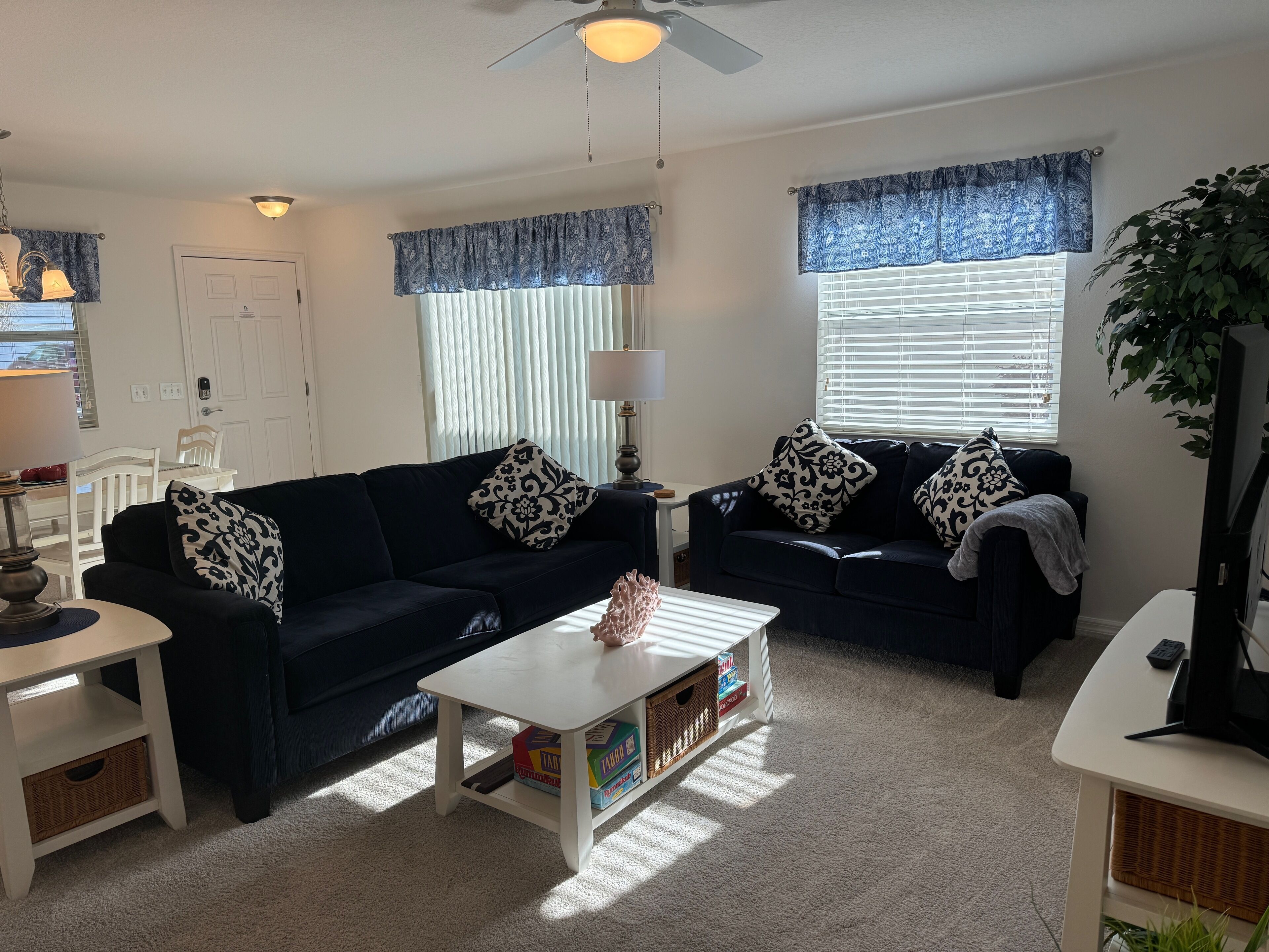 A living room with two dark sofas, a white coffee table, and a television. There are patterned throw pillows, a lamp, and a potted plant.