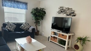Un salon avec un canapé de couleur foncée, une table basse blanche, un téléviseur posé sur un meuble blanc, une plante en pot et un décor mural représentant des poissons.