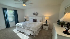 Une chambre à coucher comprenant un lit, deux tables de chevet, une commode, un ventilateur de plafond et une fenêtre avec des rideaux.