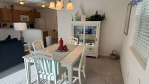 Un coin repas avec une table blanche dressée pour quatre personnes, entourée de chaises blanches. Un meuble vitré contenant des objets décoratifs est adossé au mur.