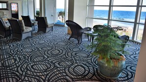 A spacious room with a patterned carpet, a large potted plant, and a view of the ocean through large windows. - Amazing Luxury Direct Ocean Front 1BD/1BA,  Modern Decor. Great Location! (Myrtle Beach)