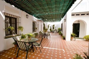 Un patio al aire libre con piso estampado, una zona de estar cubierta con sillas y una mesa, y plantas en macetas.