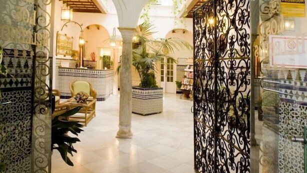 Un pasillo interior con barandillas de metal ornamentadas, suelo de baldosas y una columna central con una planta.