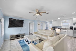 A living room with a beige sofa, a coffee table, and a TV mounted on the wall. There is a ceiling fan, a kitchen area with cabinets and a refrigerator, and a blue rug on the floor.