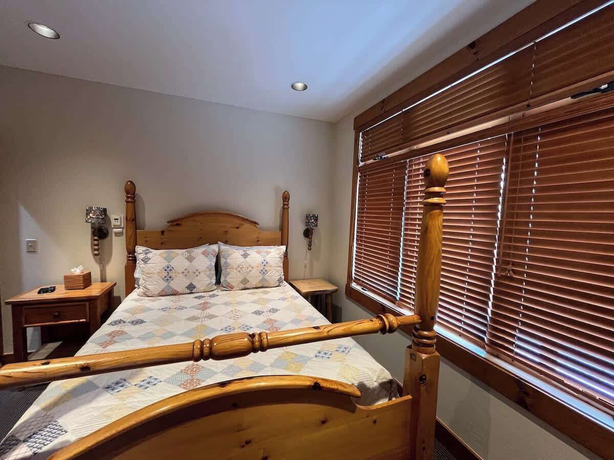 A wooden bed with a headboard, a bedside table, a lamp, and a window with blinds.