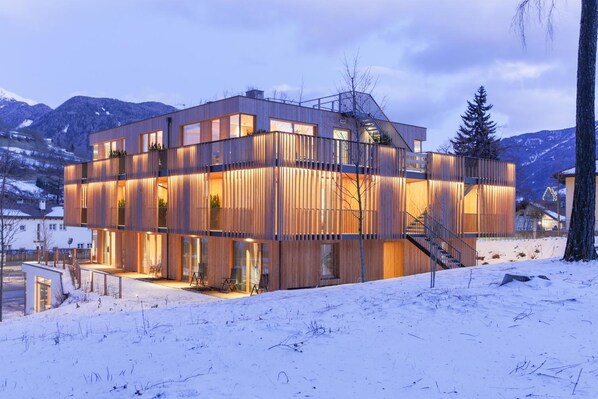 A modern multi-story building with large windows and a wooden facade, set against a snowy mountainous backdrop.