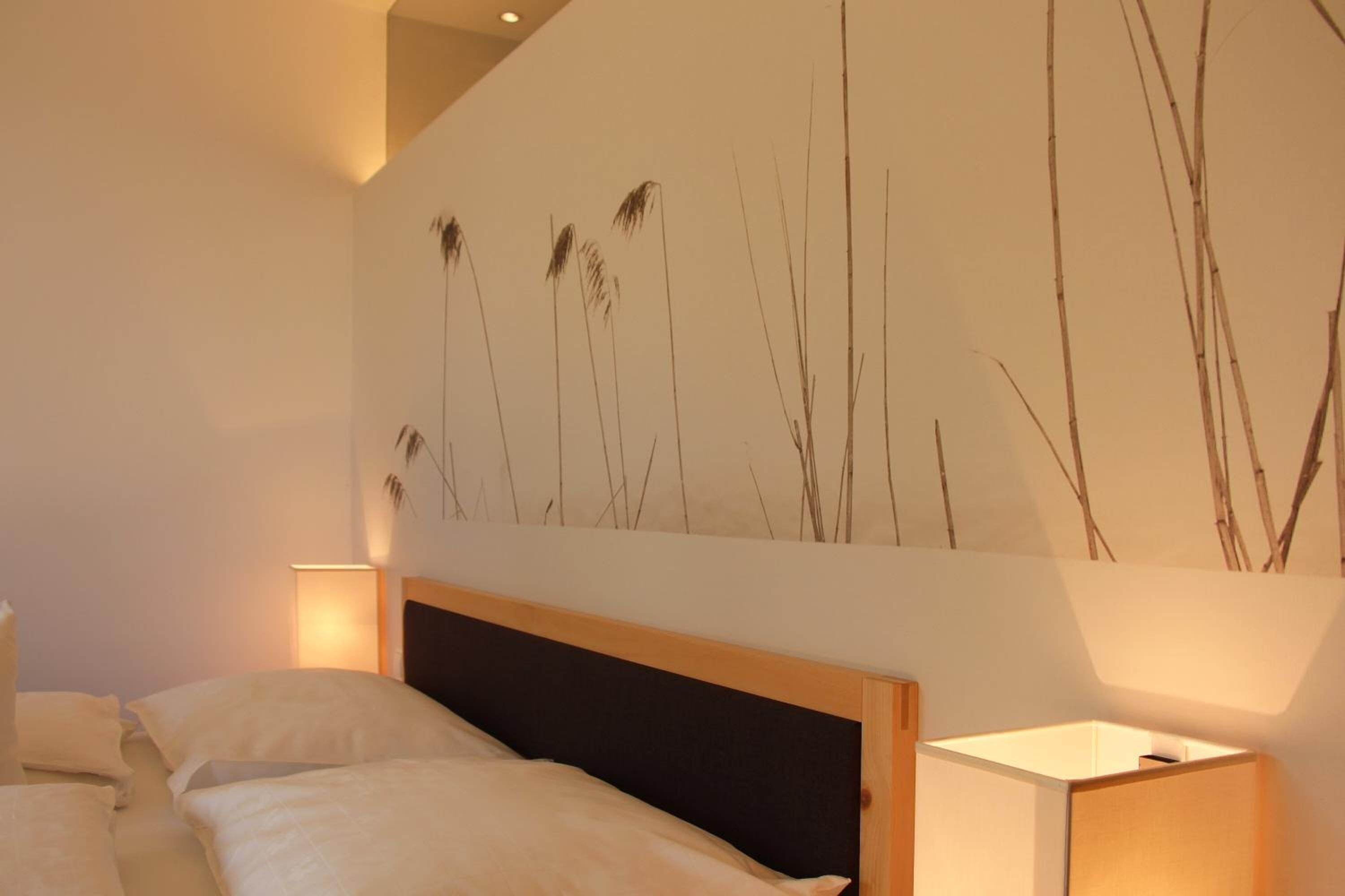 Un dormitorio con una cama, una mesita de noche, una lámpara de pared y una obra de arte que representa pastos altos.