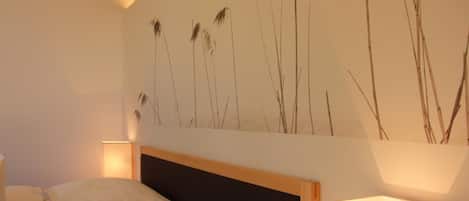 Un dormitorio con una cama, una mesita de noche, una lámpara de pared y una obra de arte que representa pastos altos.