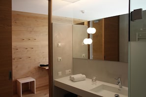 A modern bathroom with a wooden wall, a small wooden stool, a large mirror with lights, and a white sink.