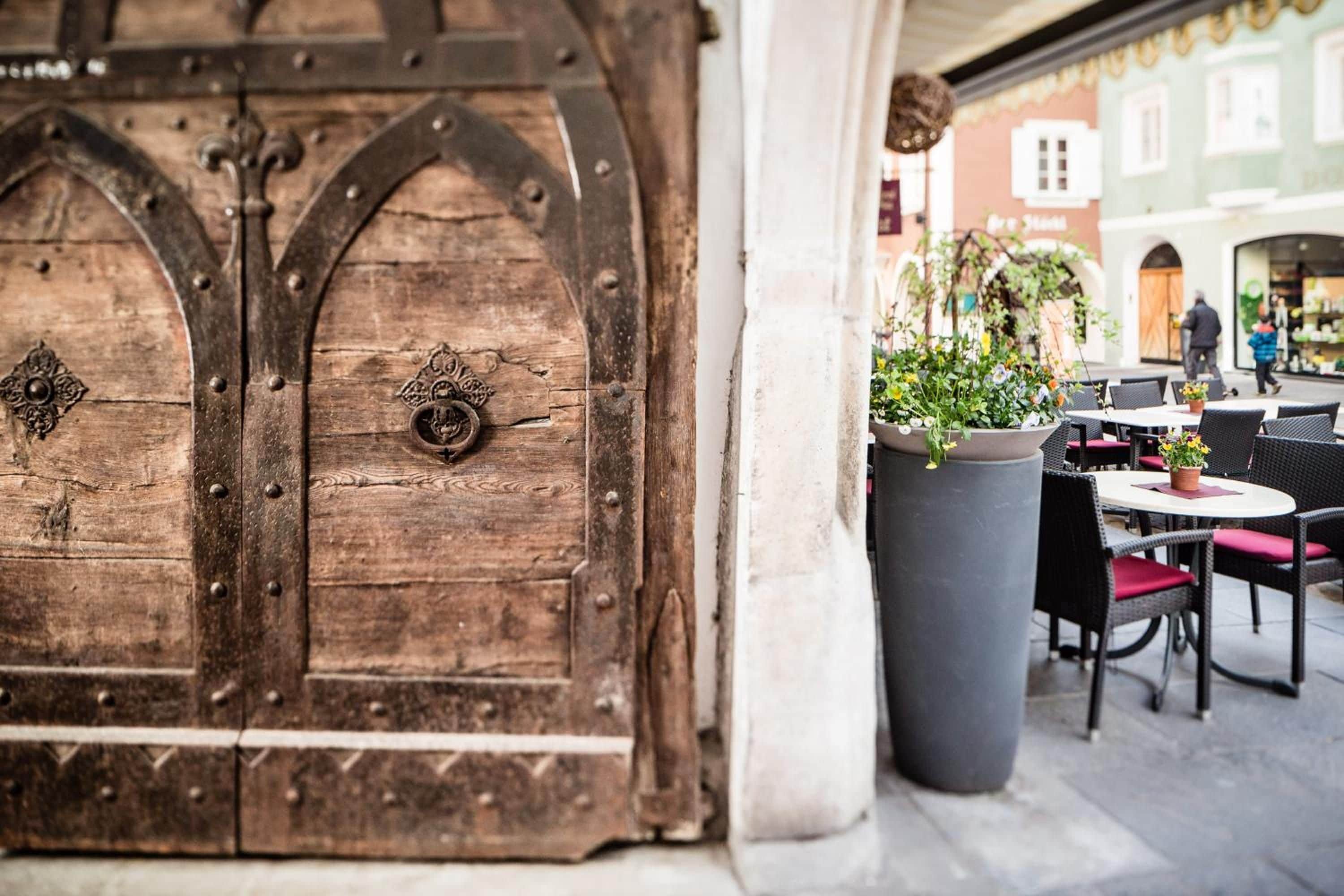 Une porte en bois rustique, dotée de charnières en métal et d’éléments décoratifs, un grand bac à fleurs et des meubles d’extérieur avec des chaises en osier.