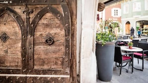 En rustik trädörr med metallgångjärn och dekorativa detaljer, en stor blomkruka och utomhusmöbler med flätade stolar.