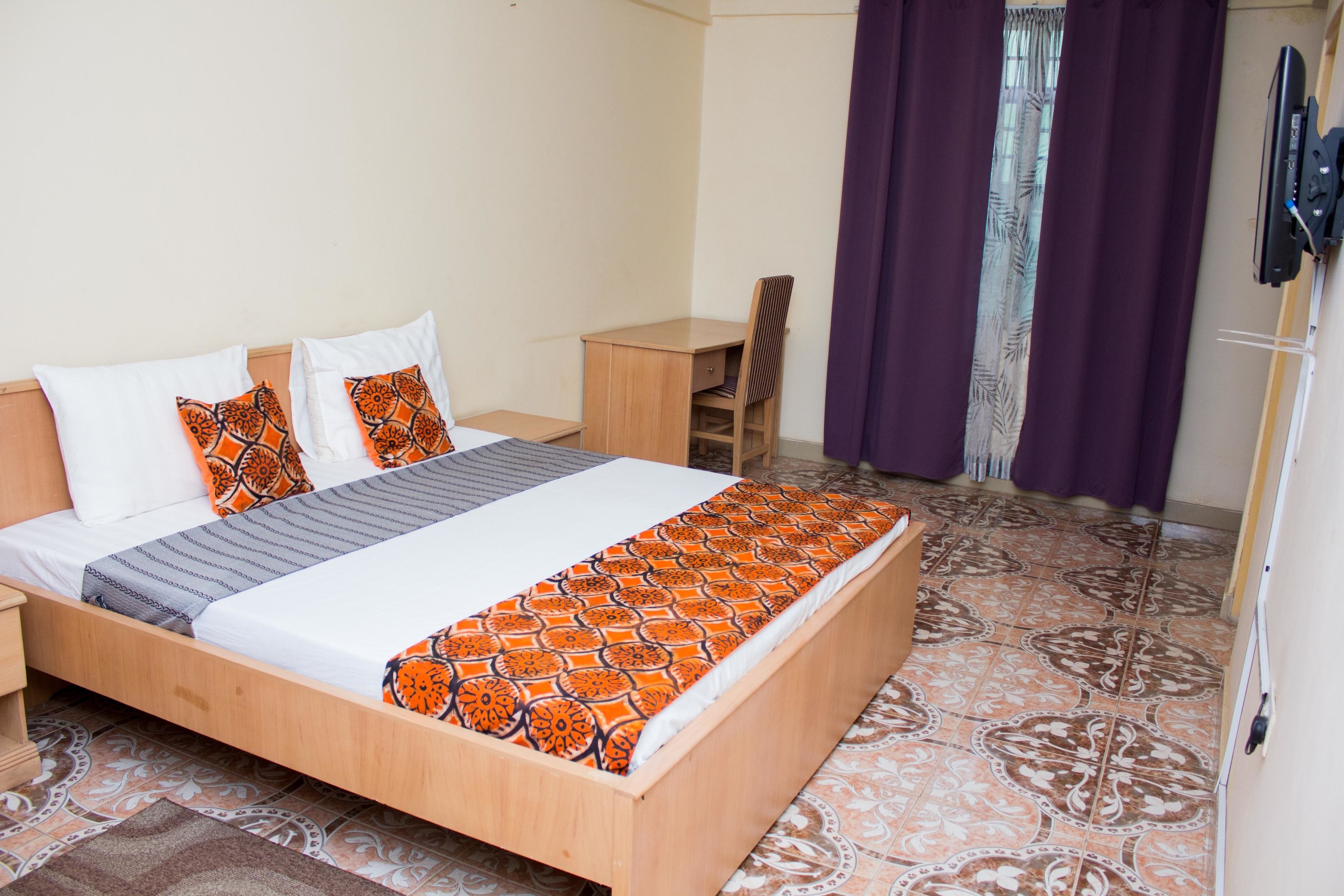A neatly made bed with patterned bedding, a wooden desk, and a chair in a room with patterned floor tiles.