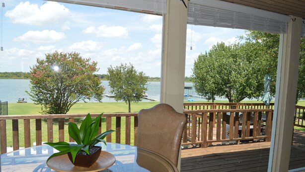 Un espace extérieur abrité, doté d’une table en verre, d’une chaise et offrant une vue sur un lac bordé d’arbres et d’une zone herbeuse.