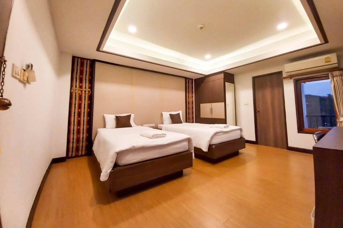 A hotel room with two beds, a wooden floor, a ceiling with recessed lighting, a window with curtains, and an air conditioning unit.