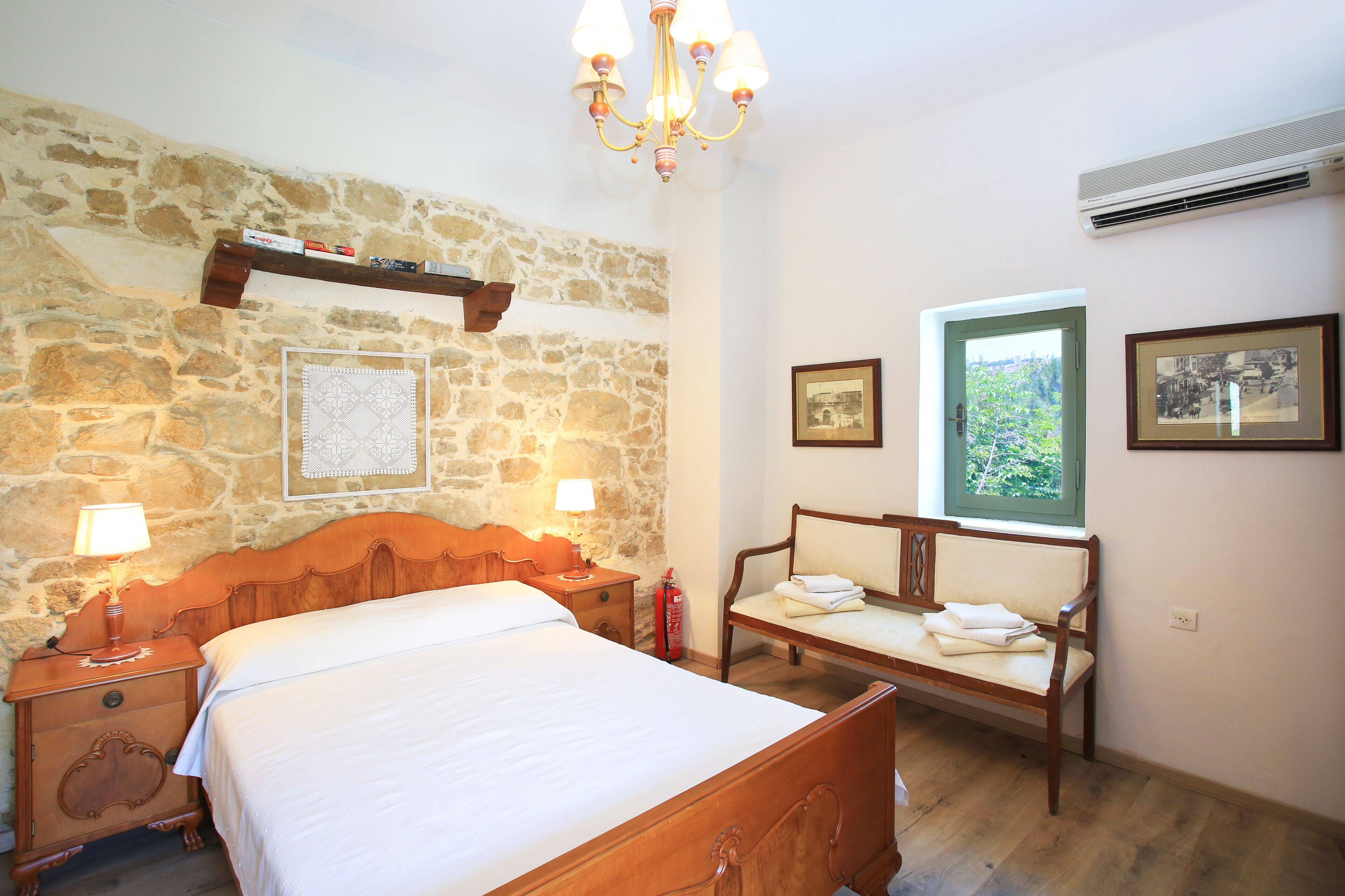 A bedroom with a stone wall, wooden bed, bedside tables with lamps, a bench with towels, and framed pictures.