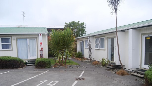 A row of single-story motel buildings with numbered parking spaces and a small garden area with palm trees and shrubs. - The Ohakune Central Backpackers & Cabins (Ohakune)