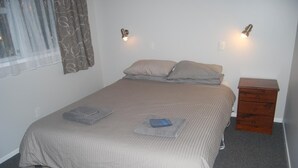 A single-bed room with a beige bedspread, two pillows, a bedside table with a lamp, and a wooden nightstand. - The Ohakune Central Backpackers & Cabins (Ohakune)