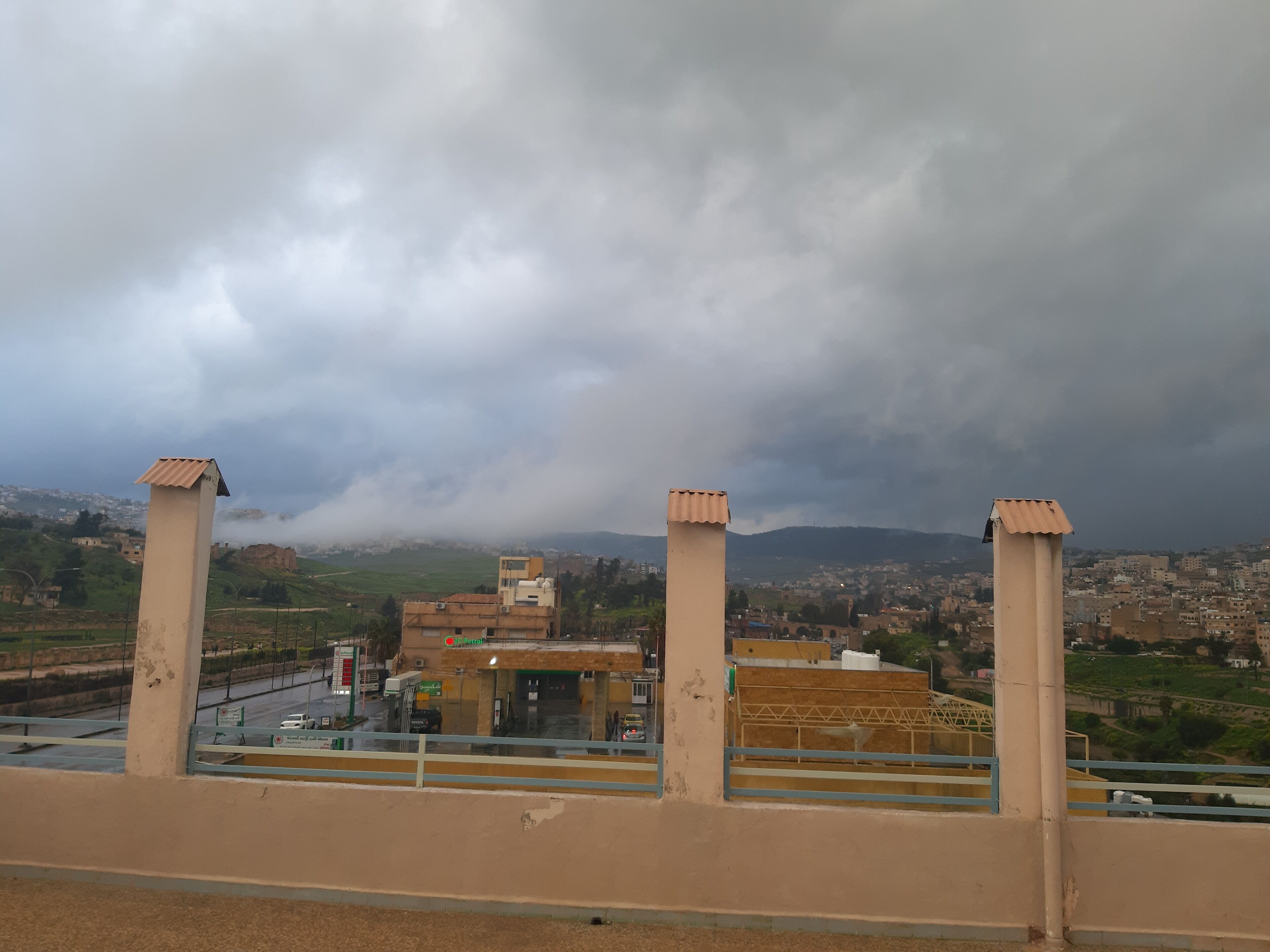 Vista de la ciudad con una gasolinera y montañas al fondo, bajo cielos nublados.