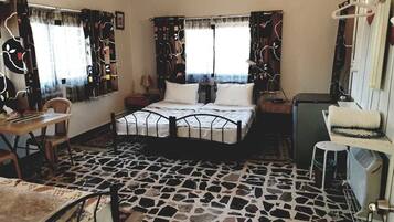 A room with a bed, a small table, and a chair. There is a window with curtains, a radiator, and a shelf with a towel.