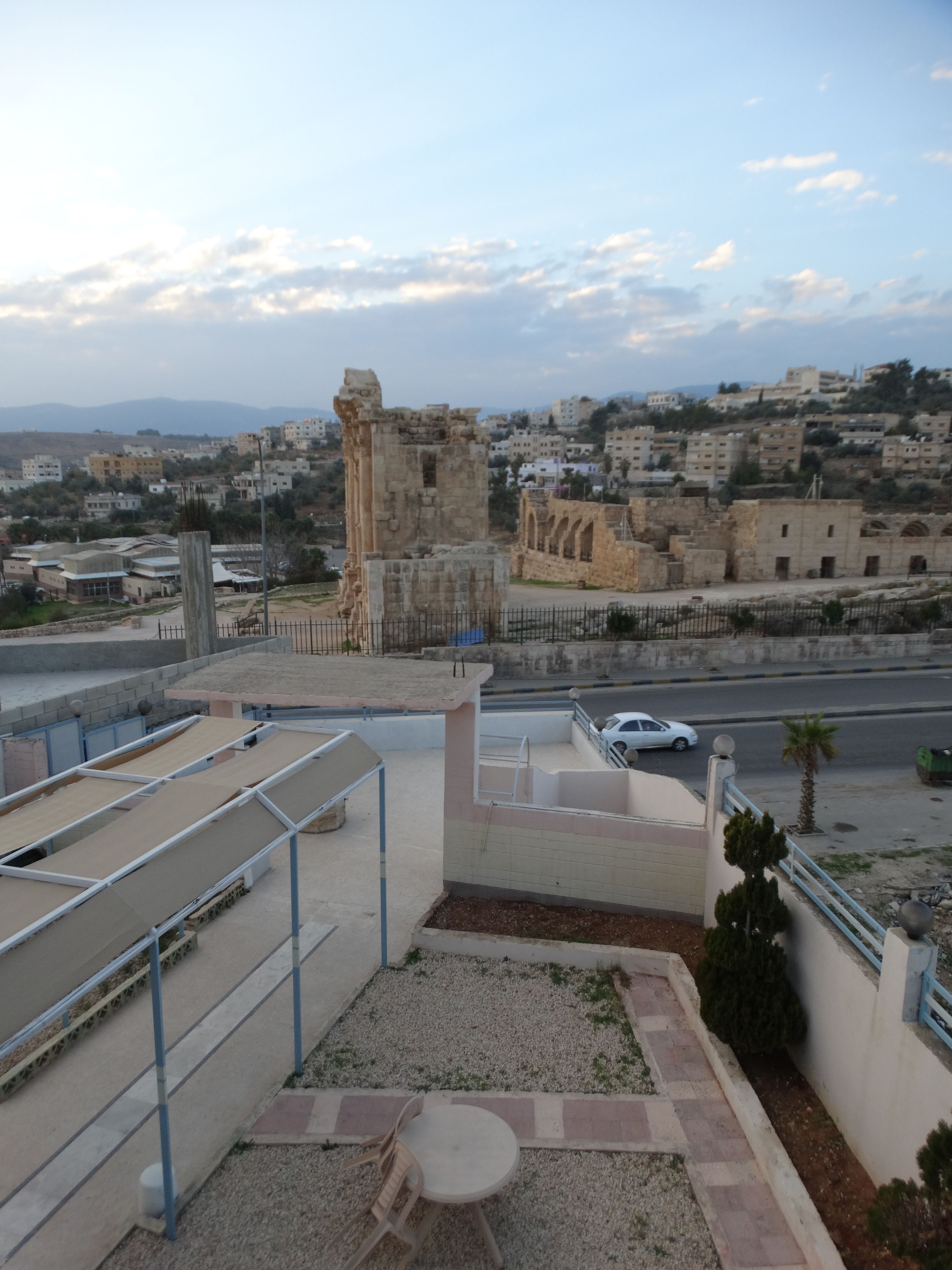 A rooftop terrace with a view of ancient ruins, a street with a parked car, and residential buildings in the background.