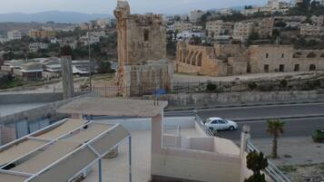 A rooftop terrace with a view of ancient ruins, a street with a parked car, and residential buildings in the background.