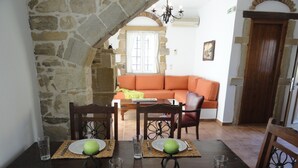Uma área de refeições com um arco de pedra, uma mesa de madeira posta com pratos, talheres e copos, e um sofá ao fundo.