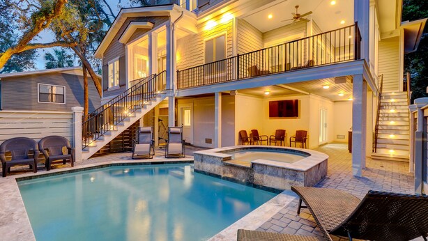 Una casa de dos pisos con piscina, mobiliario exterior y una escalera que lleva a un balcón.