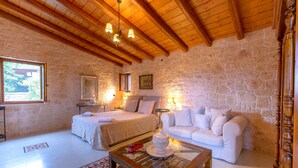 A room with a stone wall, wooden ceiling beams, a bed, a sofa, a glass coffee table, and a window with a view. - KOKKINO KASTRO is a historic estate in Aegina full of memories from 1825. (Aegina)