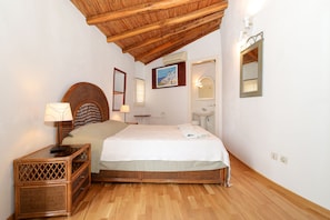 Une chambre avec un plafond en bois, un lit recouvert d’une courtepointe blanche, une table de chevet en osier, un miroir fixé au mur et un tableau encadré.