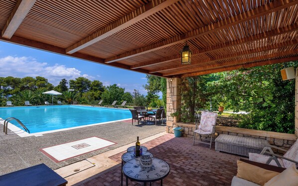 A covered outdoor pool area with lounge chairs, a table with a clock, and a view of the pool.