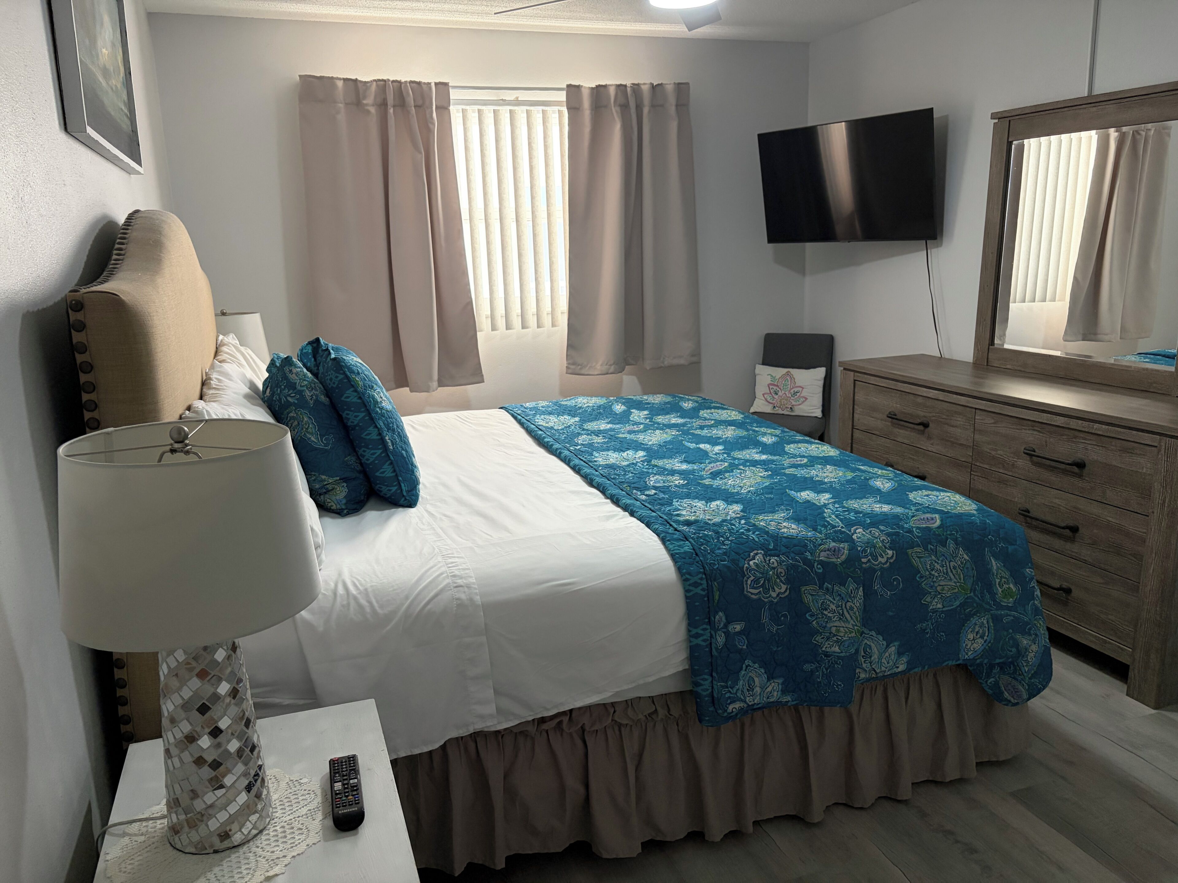 Ein Schlafzimmer mit einem Bett, einer Nachttischlampe, einem Schminktisch, einem Fernseher und einem Fenster mit Vorhängen.