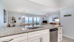 A modern kitchen with white cabinetry, a granite countertop, and a dishwasher. There is a dining area with a table and chairs, and a balcony with a view. - Award Winning Direct Gulf Front Condo with a Fantastic Sunset View (Indian Shores)