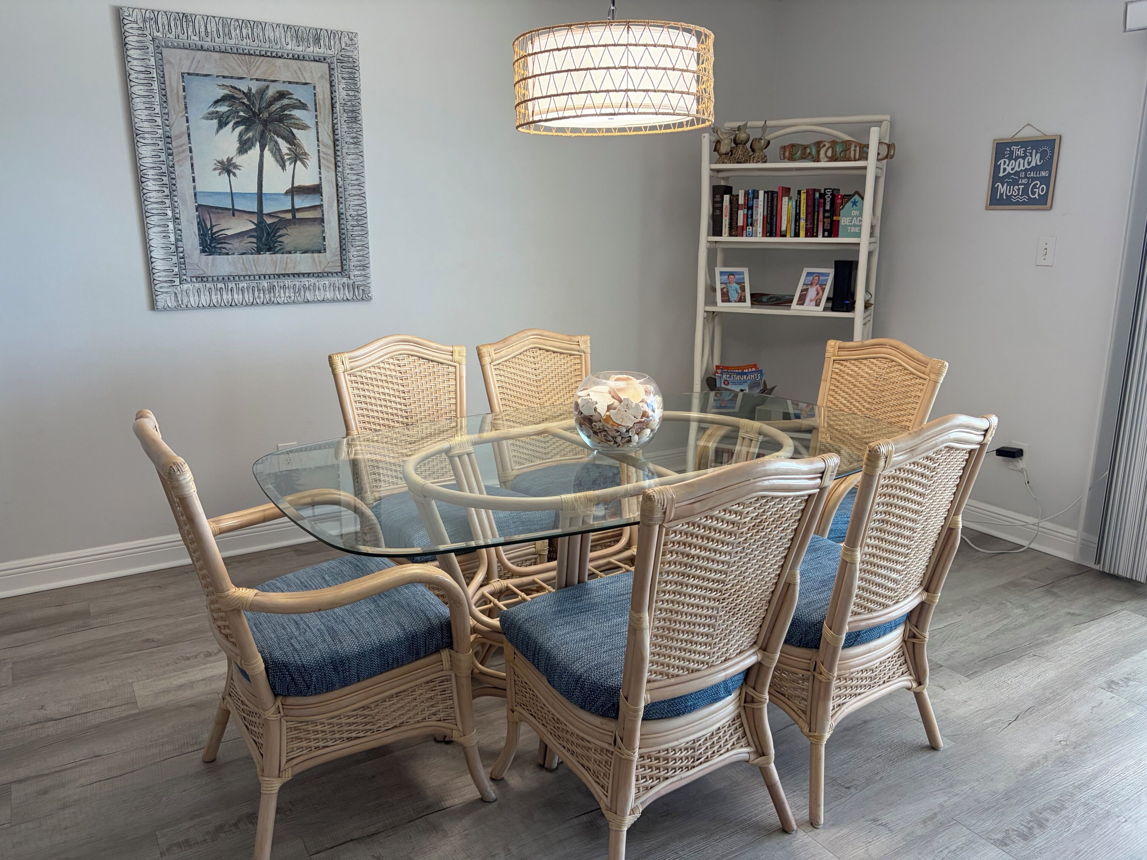 Un coin repas avec une table en verre entourée de chaises en osier dotées de coussins bleus, une décoration murale et une bibliothèque contenant des livres et une photographie encadrée.