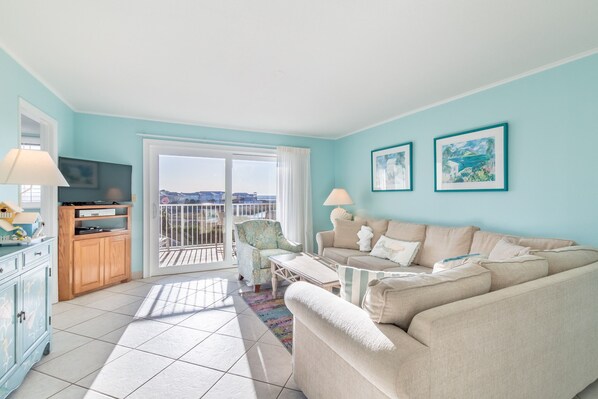 A living room with a beige sofa, a small table, a chair, and a balcony with a view.