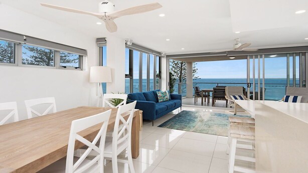 Uma sala de estar moderna com uma mesa de jantar de madeira, cadeiras brancas, um sofá azul e vista para o oceano.