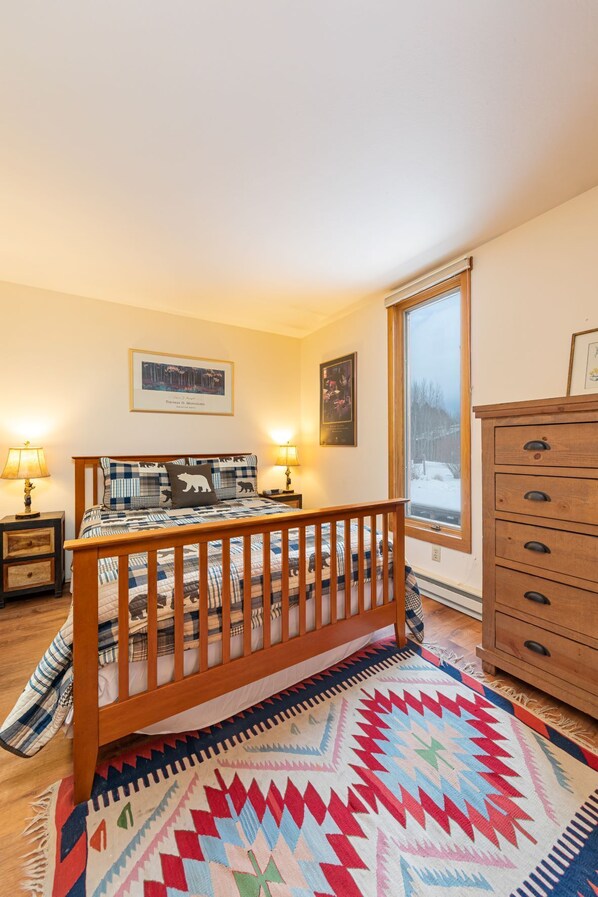 A bedroom with a wooden bed, two bedside tables with lamps, a dresser, and a large window with a view of a snowy landscape.