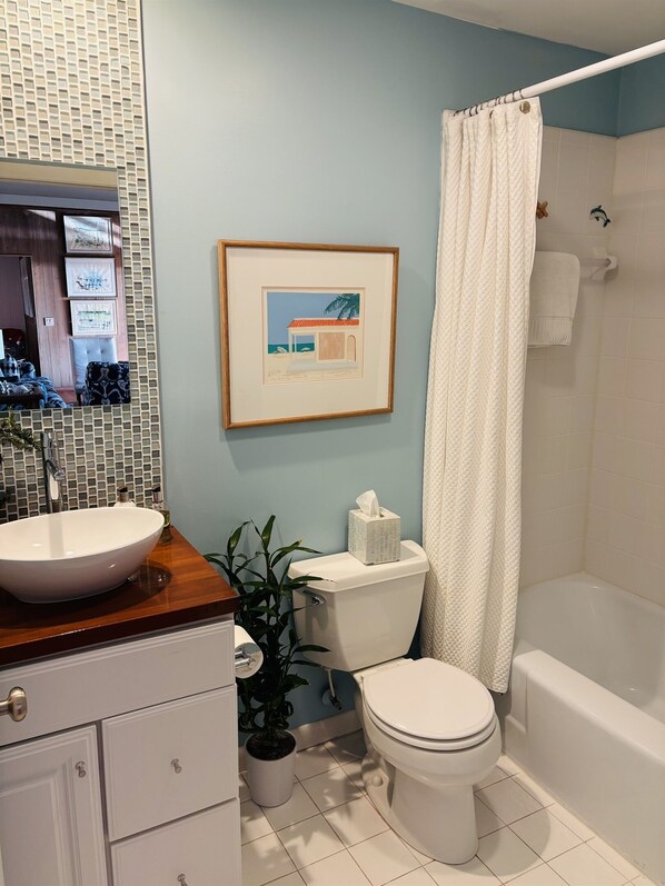 A bathroom with a white sink, toilet, and bathtub. There is a framed picture on the wall and a potted plant near the toilet.