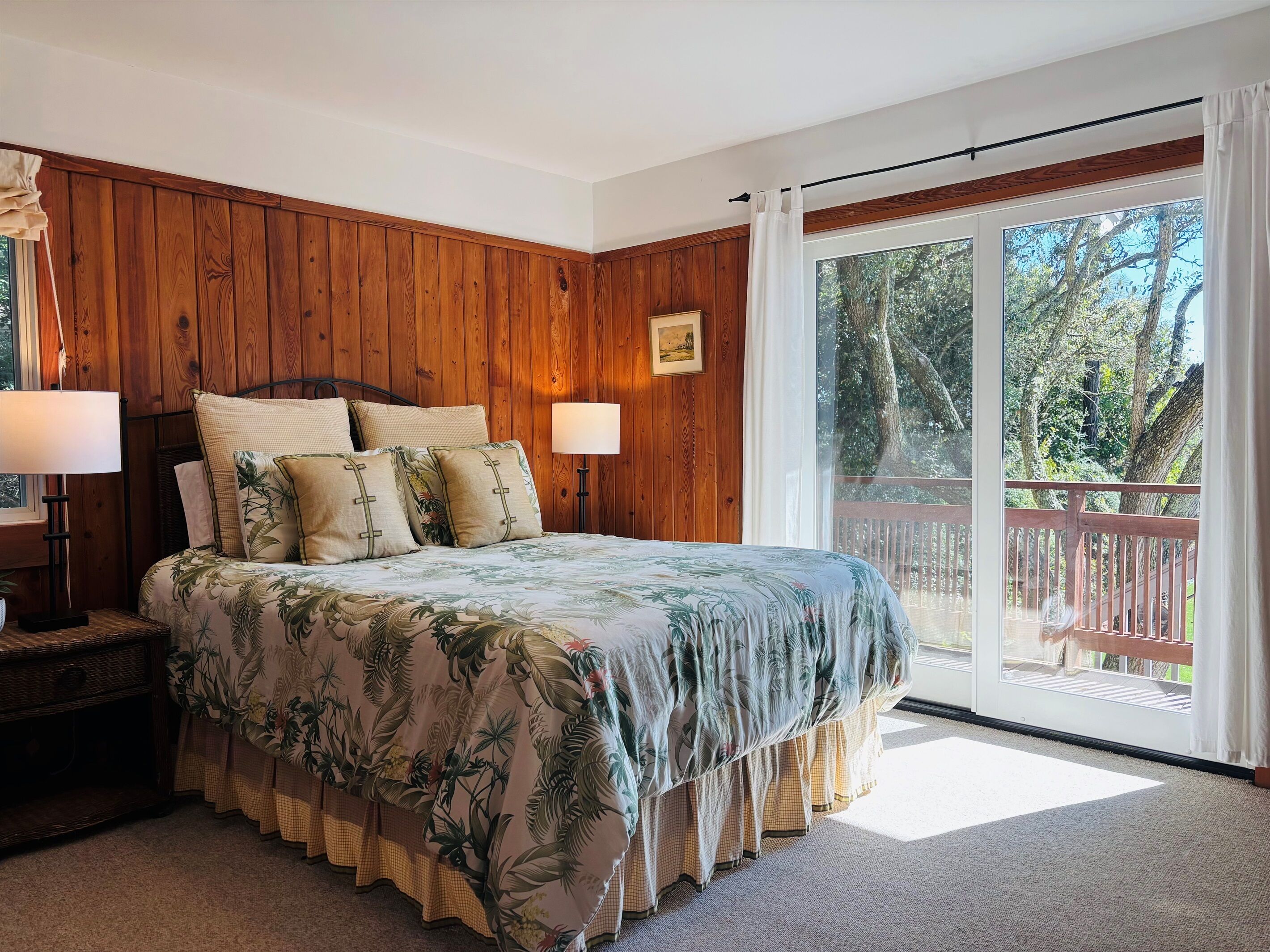 A bedroom with a wooden paneled wall, a large bed with floral bedding, two bedside lamps, and a view of a deck through a sliding glass door.