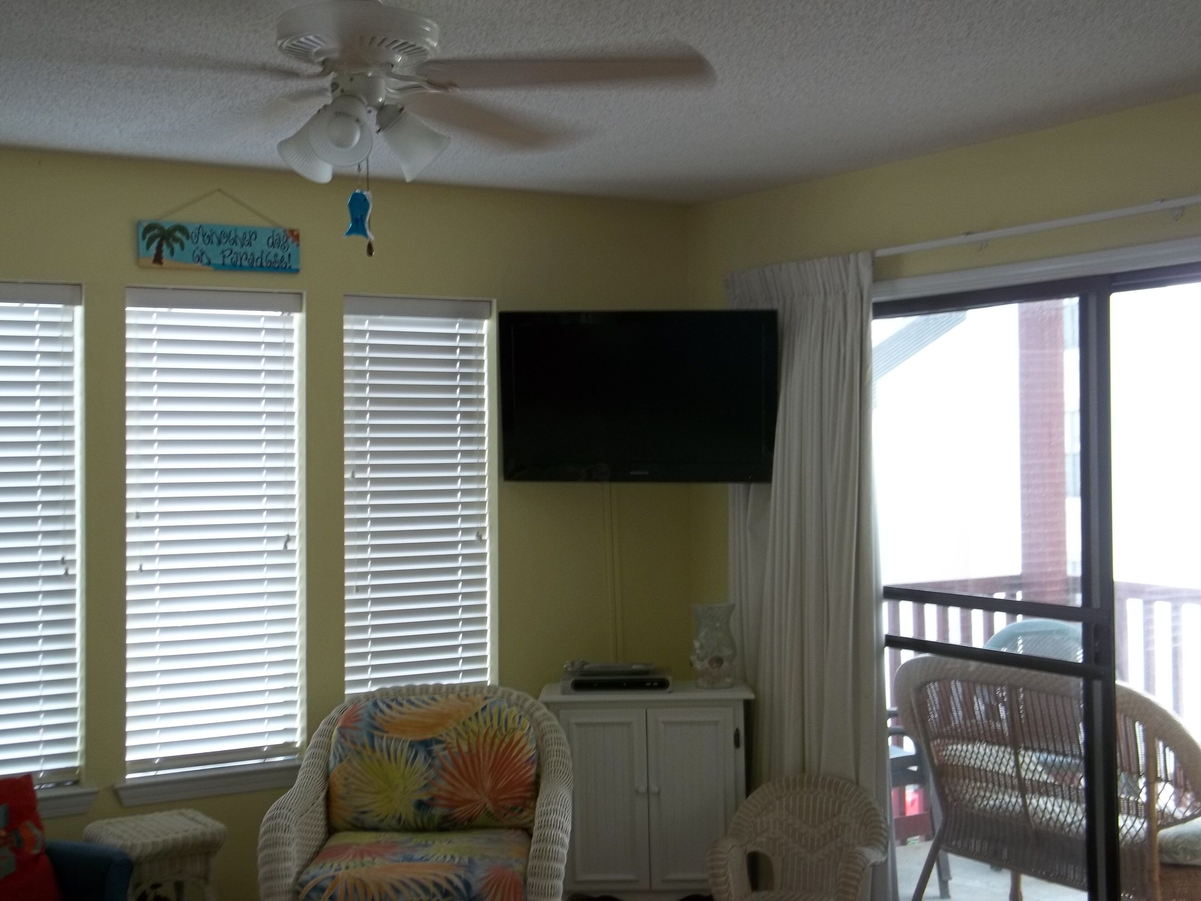 A room with a ceiling fan, a television mounted on the wall, a window with blinds, a wicker chair, and a view of the outside.