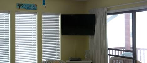 A room with a ceiling fan, a television mounted on the wall, a window with blinds, a wicker chair, and a view of the outside.