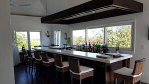 Eine Küche mit Barbereich, Korbstühlen an der Bar, einem großen Fenster mit Ausblick und einer hängenden Leuchte.