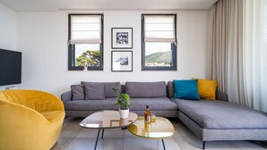 Una sala moderna con un sofá seccional gris, un sillón amarillo y una mesa de centro de vidrio. Hay dos ventanas con cortinas blancas, un televisor de pantalla plana a la izquierda y una planta en maceta sobre el sofá.
